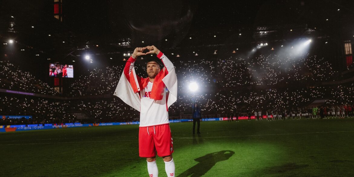Lukas Podolski stands in the spotlight at the RheinEnergieStadion wearing the 1. FC Köln jersey and forms a heart with his hands.