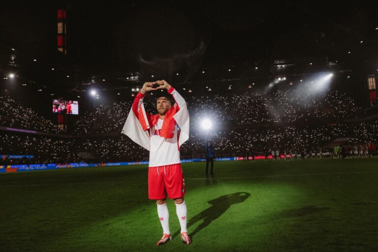 Lukas Podolski stands in the spotlight at the RheinEnergieStadion wearing the 1. FC Köln jersey and forms a heart with his hands.