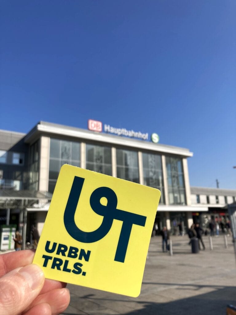 Eine Hand hält einen gelben Aufkleber mit einer grünen Aufschrift "UT | URBN TRLS." in die Linse. Dahinter ist der Dortmunder Hauptbahnhof zu erkennen.