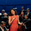 Eine junge Frau in einem hellroten Abendkleid steht vor einem Orchester, ihr Cello in einer Hand, und schaut ergriffen Richtung Kamera ins Publikum.