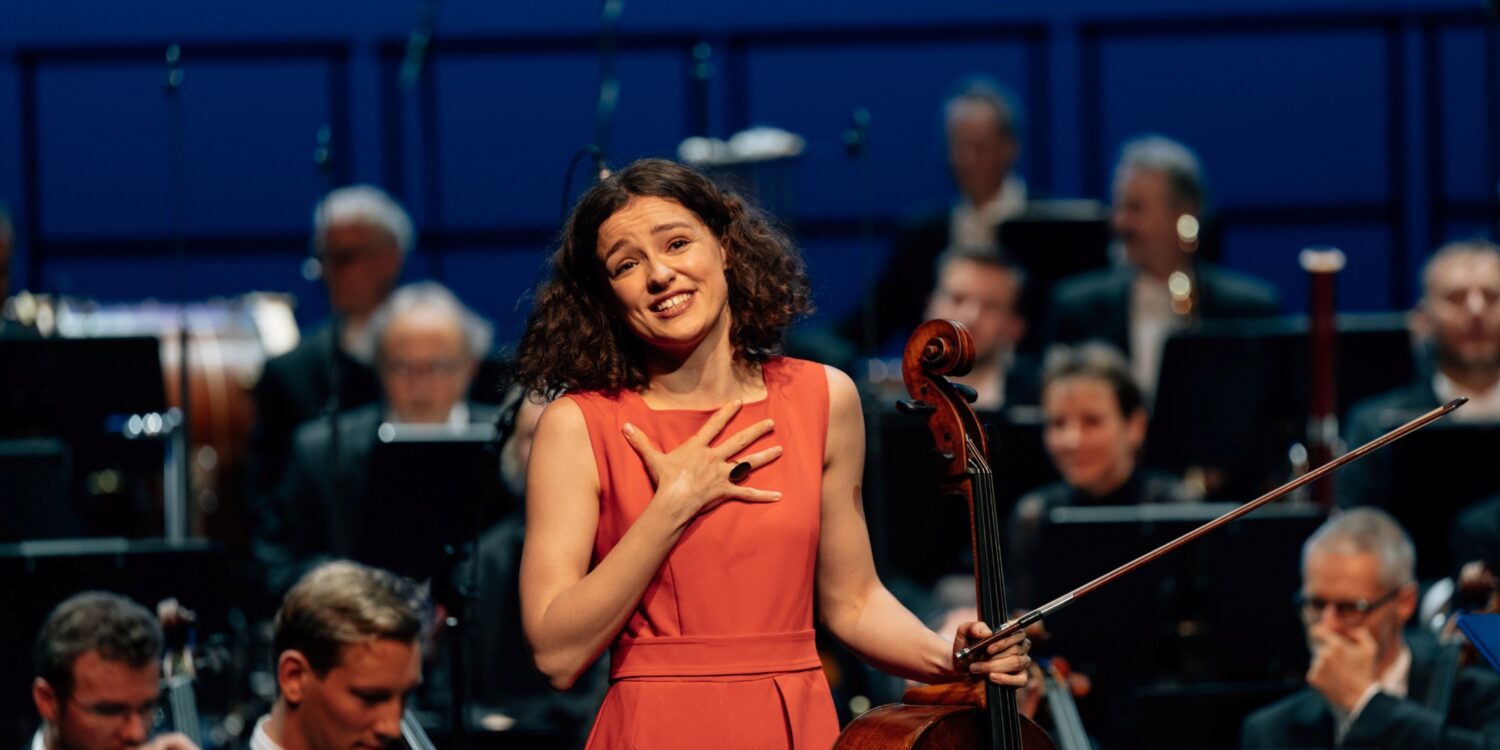 Eine junge Frau in einem hellroten Abendkleid steht vor einem Orchester, ihr Cello in einer Hand, und schaut ergriffen Richtung Kamera ins Publikum.