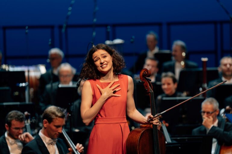 Eine junge Frau in einem hellroten Abendkleid steht vor einem Orchester, ihr Cello in einer Hand, und schaut ergriffen Richtung Kamera ins Publikum.