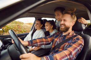 A man, a woman, and two children are sitting in a car; the man is behind the wheel. They are clearly having fun; the wind is blowing the man’s hair around through the open window.