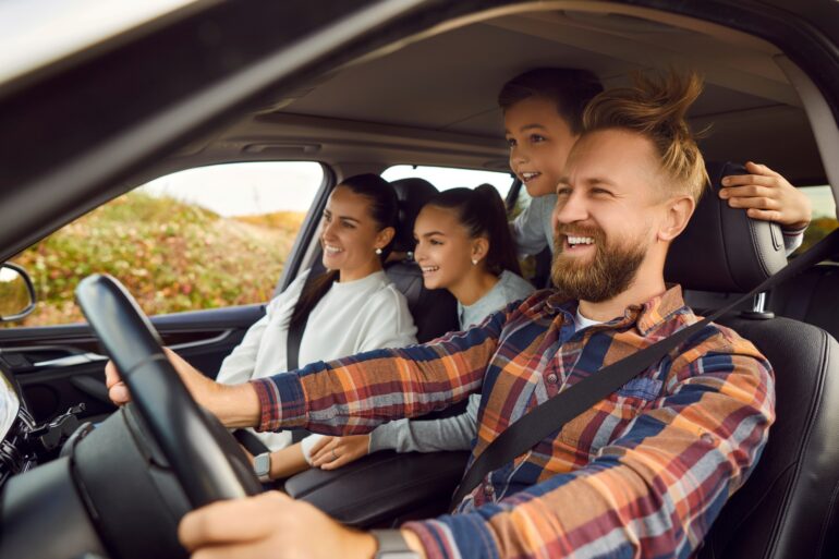 A man, a woman, and two children are sitting in a car; the man is behind the wheel. They are clearly having fun; the wind is blowing the man’s hair around through the open window.