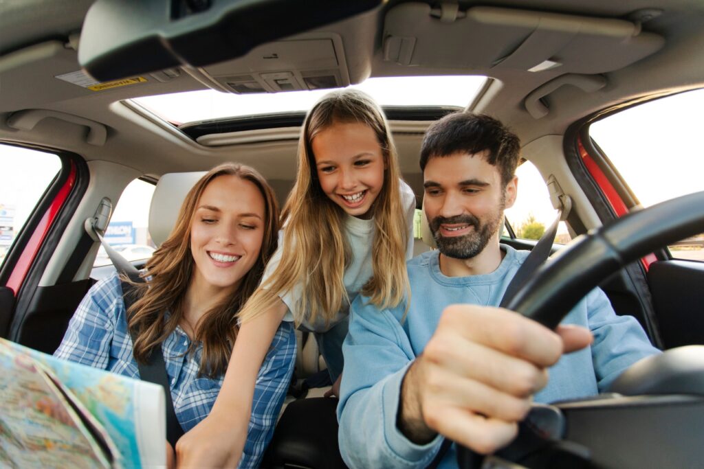 A man and a woman are sitting in a car; a child is leaning forward from the back seat between them. All three are looking at a map that the woman in the passenger seat is holding.