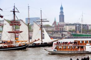 Zwei historische Segelschiffe und ein Ausflugsdampfer fahren auf der Elbe in Hamburg. Das Ufer im Hintergrund ist voll mit Zuschauern. Dahinter ragt der Hamburger Michel aus der Stadtkulisse auf.