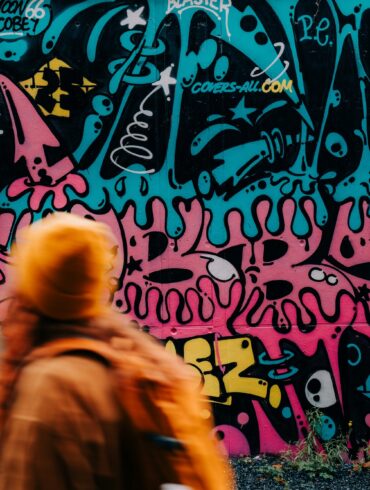 A woman wearing an orange hat and jacket is running quickly past a wall covered in teal-and-pink graffiti.