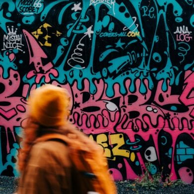 A woman wearing an orange hat and jacket is running quickly past a wall covered in teal-and-pink graffiti.
