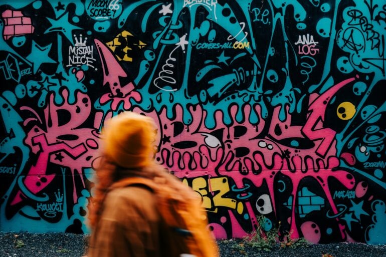 A woman wearing an orange hat and jacket is running quickly past a wall covered in teal-and-pink graffiti.