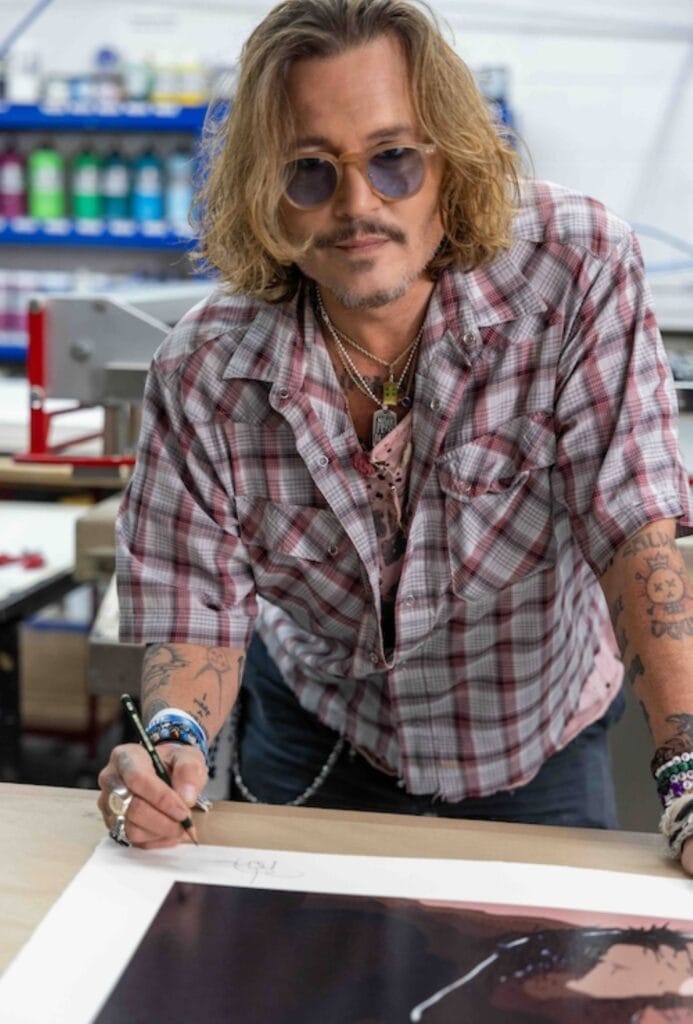 Der Schauspieler Johnny Depp beugt sich über einen Tisch, um ein Bild zu signieren. Dabei lächelt er in die Kamera.