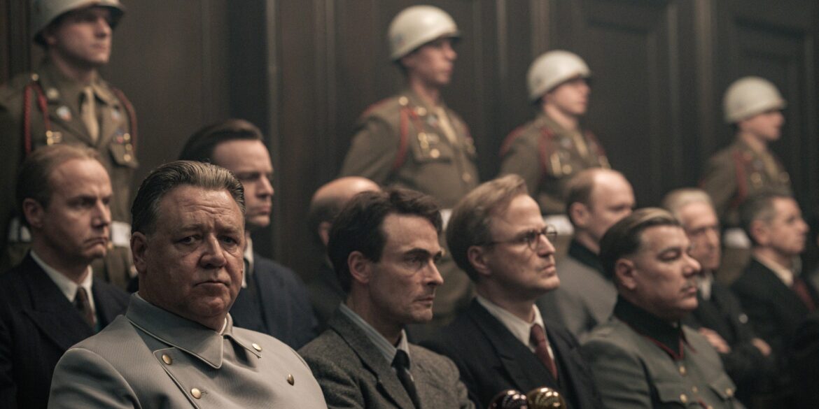 A still from the film “Nuremberg” (to be released in May in Germany). Several people in Nazi uniforms are seated in the dock, with U.S. soldiers wearing white helmets standing behind them. Seated at the front of the dock is actor Russell Crowe as Hermann Göring, who glances briefly toward the camera.