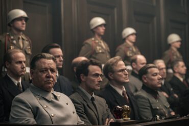 A still from the film “Nuremberg” (to be released in May in Germany). Several people in Nazi uniforms are seated in the dock, with U.S. soldiers wearing white helmets standing behind them. Seated at the front of the dock is actor Russell Crowe as Hermann Göring, who glances briefly toward the camera.
