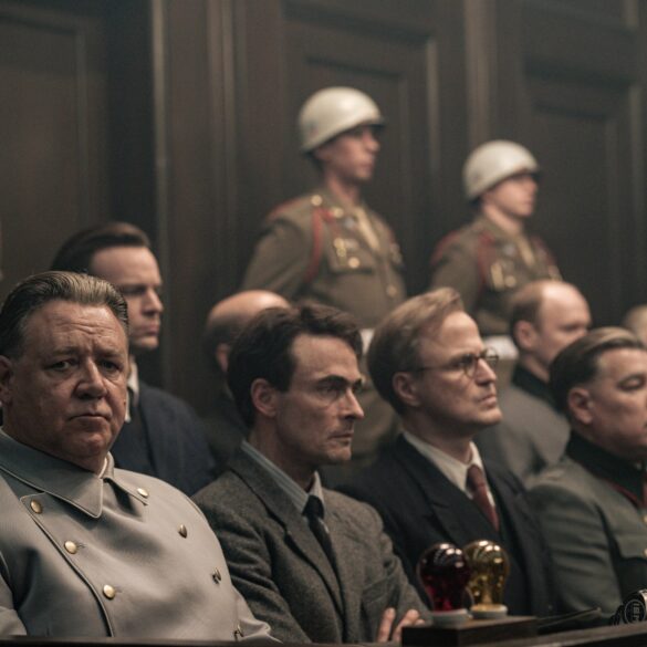 A still from the film “Nuremberg” (to be released in May in Germany). Several people in Nazi uniforms are seated in the dock, with U.S. soldiers wearing white helmets standing behind them. Seated at the front of the dock is actor Russell Crowe as Hermann Göring, who glances briefly toward the camera.