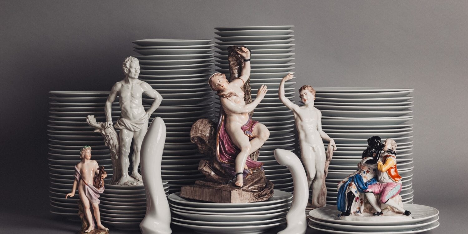 Vor mehreren Stapeln Porzellanteller sind mehrere Porzellanfiguren aufgestellt, die unterschiedliche Menschen darstellen, aber immer nackt oder leicht bekleidet. Dazwischen zwei Porzellanfiguren in Form von Sexspielzeug.