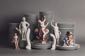 Vor mehreren Stapeln Porzellanteller sind mehrere Porzellanfiguren aufgestellt, die unterschiedliche Menschen darstellen, aber immer nackt oder leicht bekleidet. Dazwischen zwei Porzellanfiguren in Form von Sexspielzeug.