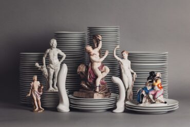 Vor mehreren Stapeln Porzellanteller sind mehrere Porzellanfiguren aufgestellt, die unterschiedliche Menschen darstellen, aber immer nackt oder leicht bekleidet. Dazwischen zwei Porzellanfiguren in Form von Sexspielzeug.