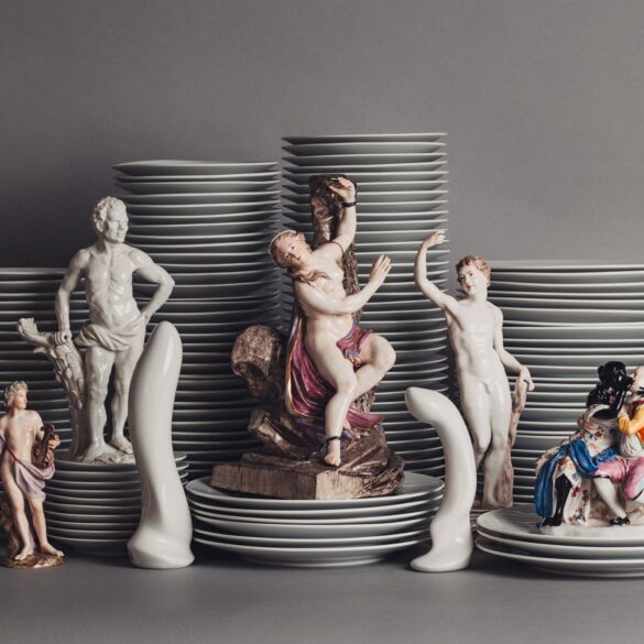 Vor mehreren Stapeln Porzellanteller sind mehrere Porzellanfiguren aufgestellt, die unterschiedliche Menschen darstellen, aber immer nackt oder leicht bekleidet. Dazwischen zwei Porzellanfiguren in Form von Sexspielzeug.