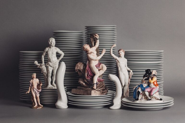 Vor mehreren Stapeln Porzellanteller sind mehrere Porzellanfiguren aufgestellt, die unterschiedliche Menschen darstellen, aber immer nackt oder leicht bekleidet. Dazwischen zwei Porzellanfiguren in Form von Sexspielzeug.