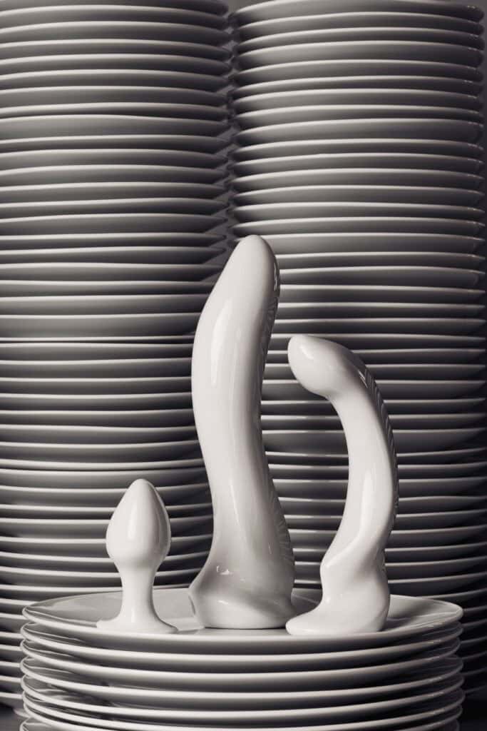 Auf einem Stapel Porzellanteller in einer Ausstellung im Museum Schloss Fürstenberg stehen drei Sextoys, ebenfalls aus Porzellan.