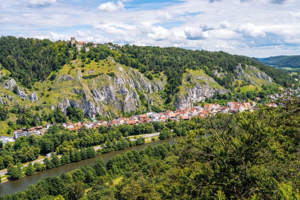 Über einem grünen Flusstal mit einer langezogenen Stadt erhebt sich eine Burgruine auf einem Felsvorsprung, umgeben von sommergrünem Wald.