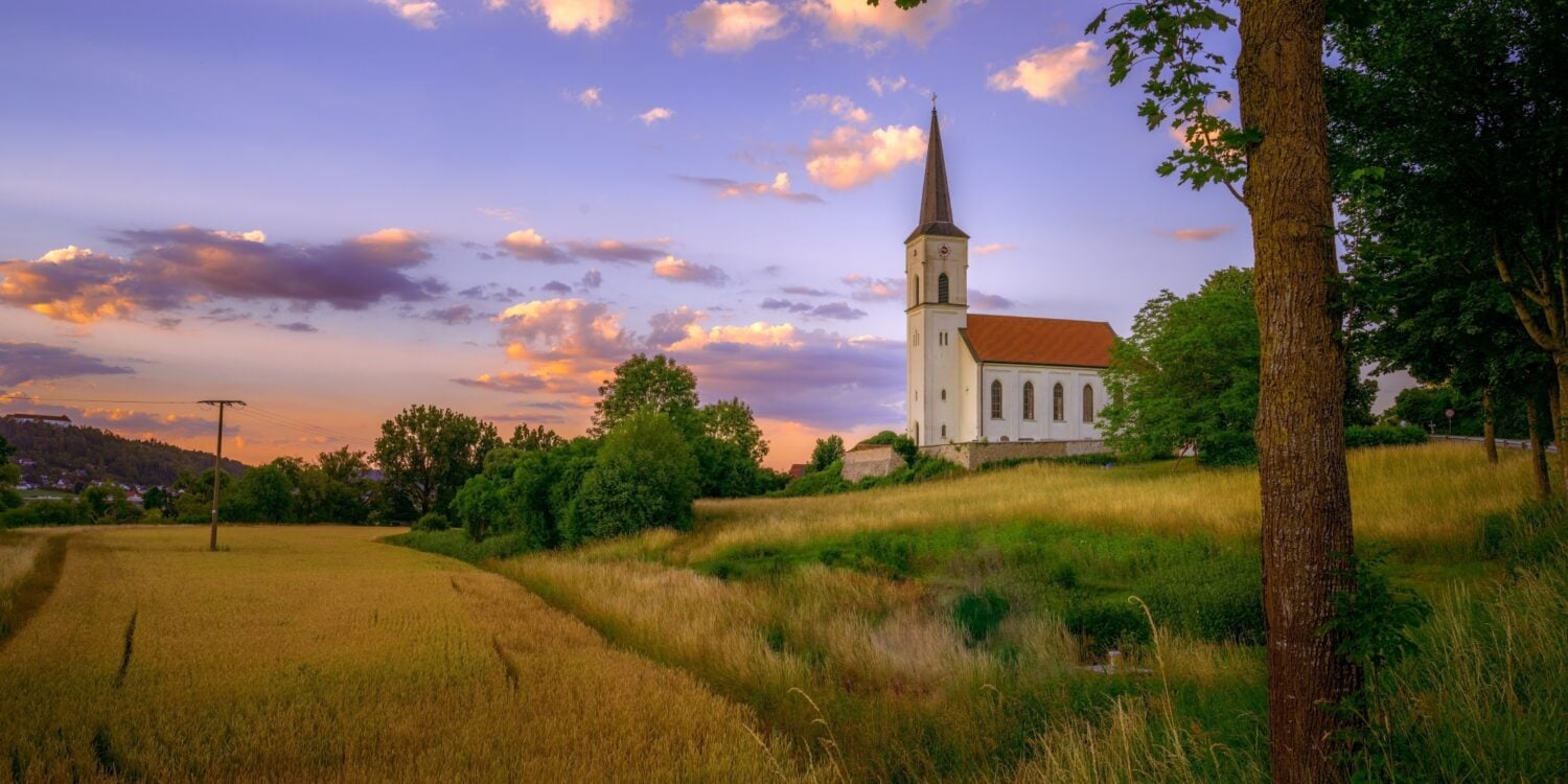 In einem korngelben Feld steht zur rechten eine kleine, weiße Kirche mit rotem Dach. Der Himmel darüber ist im Sonnenuntergang in lila und orange gefärbt.
