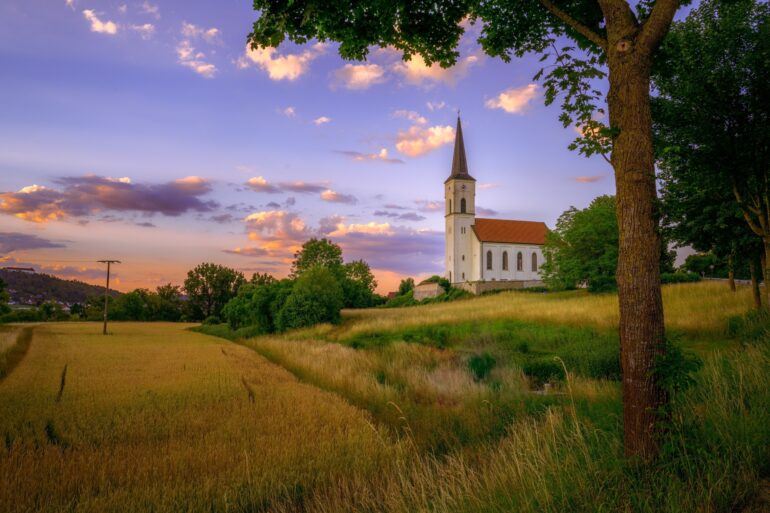 In einem korngelben Feld steht zur rechten eine kleine, weiße Kirche mit rotem Dach. Der Himmel darüber ist im Sonnenuntergang in lila und orange gefärbt.
