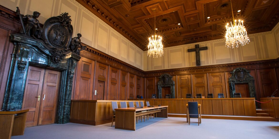 Ein historischer Gerichtssaal mit dunkler Holzvertäfelung an den Wänden. Über der Richterbank hängt ein großes, hölzernes Kreuz, die Eingangstür links wird von einem breiten Portal aus dunklem Marmor umrahmt.