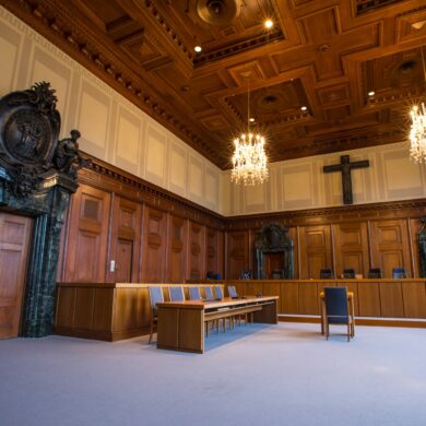 Ein historischer Gerichtssaal mit dunkler Holzvertäfelung an den Wänden. Über der Richterbank hängt ein großes, hölzernes Kreuz, die Eingangstür links wird von einem breiten Portal aus dunklem Marmor umrahmt.