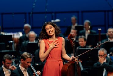 A young woman in a bright red evening gown stands in front of an orchestra, her cello in one hand, and looks, visibly moved, toward the camera and into the audience.