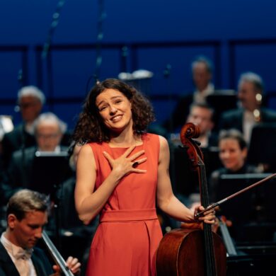A young woman in a bright red evening gown stands in front of an orchestra, her cello in one hand, and looks, visibly moved, toward the camera and into the audience.
