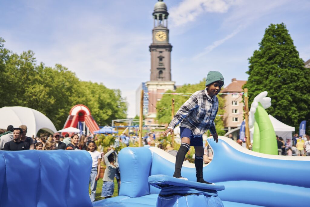 A child is surfing on a rubber surfboard in a world of rubber waves, with people at a fair in the background. A bell tower can be seen in the background.