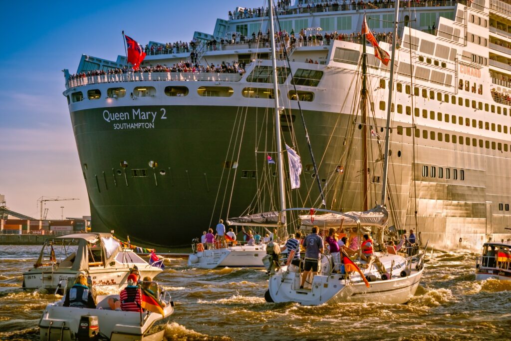 Several small motorboats are sailing in a harbor basin in front of the cruise ship “Queen Mary 2.”