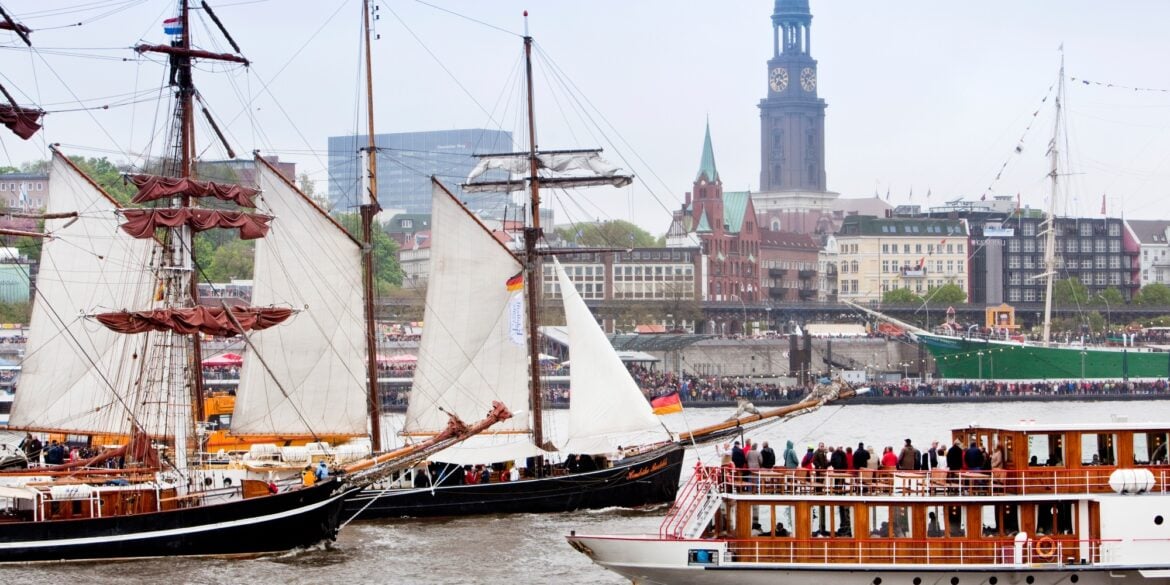 Two historic sailing ships and a sightseeing steamboat are sailing on the Elbe River in Hamburg. The riverbank in the background is crowded with spectators. Behind them, Hamburg’s St. Michael’s Church rises above the city skyline.