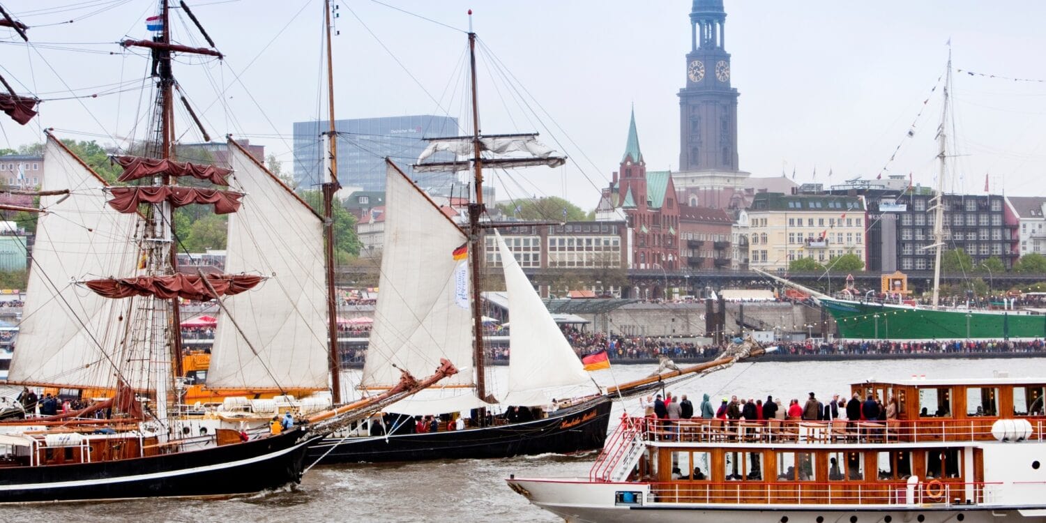 Two historic sailing ships and a sightseeing steamboat are sailing on the Elbe River in Hamburg. The riverbank in the background is crowded with spectators. Behind them, Hamburg’s St. Michael’s Church rises above the city skyline.