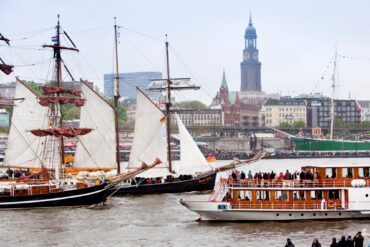 Two historic sailing ships and a sightseeing steamboat are sailing on the Elbe River in Hamburg. The riverbank in the background is crowded with spectators. Behind them, Hamburg’s St. Michael’s Church rises above the city skyline.