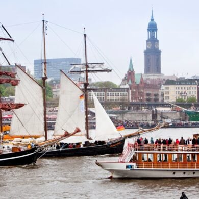 Two historic sailing ships and a sightseeing steamboat are sailing on the Elbe River in Hamburg. The riverbank in the background is crowded with spectators. Behind them, Hamburg’s St. Michael’s Church rises above the city skyline.