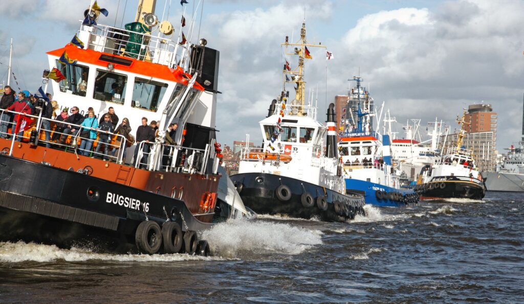 Four tugboats, fully loaded with passengers, are leaving the Hamburg harbor basin in single file.