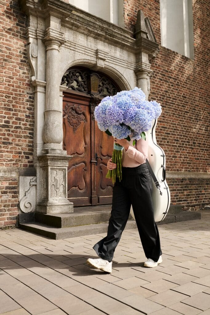 Eine Person mit einem Cello-Koffer auf dem Rücken geht vor einer Doppelholztür an einem Ziegelgebäude vorbei. In der Hand hält sie einen riesigen Strauch mit Blumen, die ihr Gesicht verdecken.