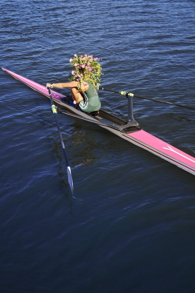 Eine Person rudert in einem rosa Sportkanu auf dem Wasser. Über der Schulter trägt sie einen riesigen Blumenstrauß, der ihr Gesicht verdeckt.