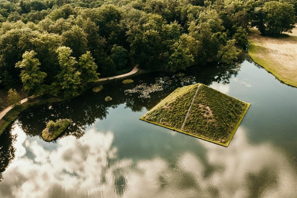 Drohnenaufnahme eines grünen Schlossparks mit See. Mitten im See ragt eine überwachsene, künstliche Pyramide aus dem Wasser.