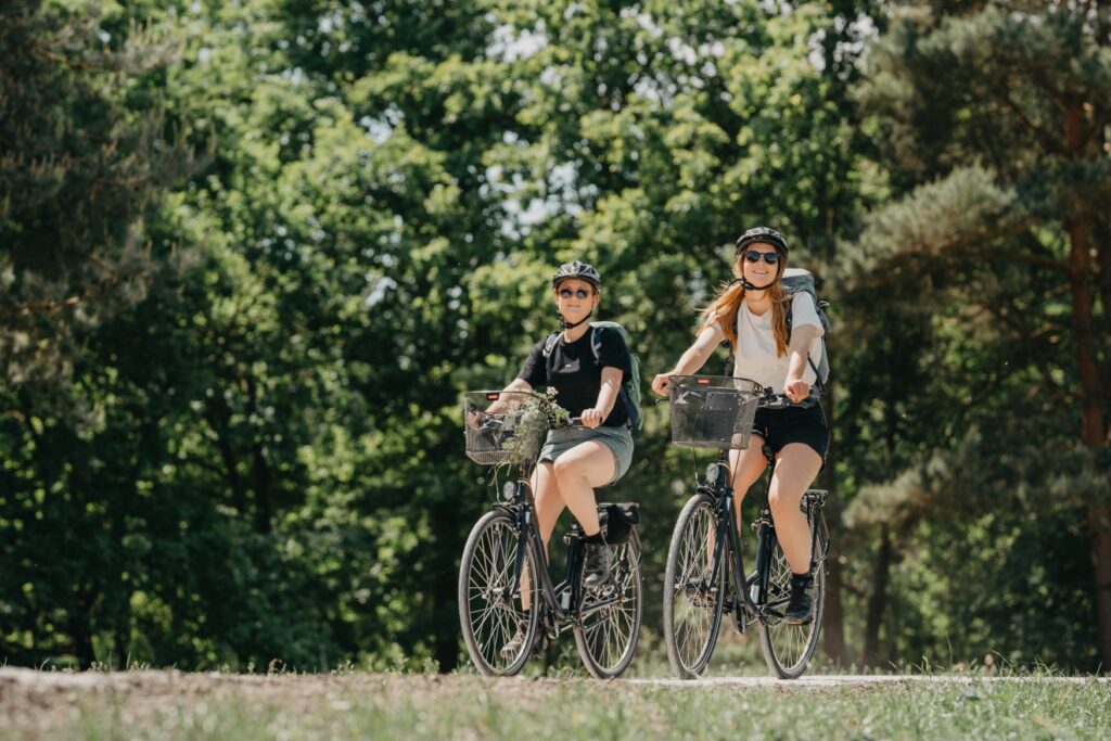 Zwei junge Frauen in sommerlicher Kleidung fahren auf Fahrrädern über eine Lichtung, hinter ihnen grüner Wald.