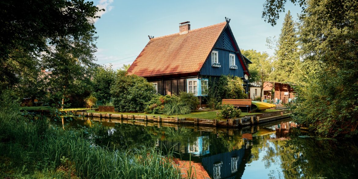 Ein schwarzes Haus mit rotem Dach steht in der Biege einer Flussschleife. Am Flussufer sind mehrere Kanus aufgebockt. Der Himmel über allem ist blau.