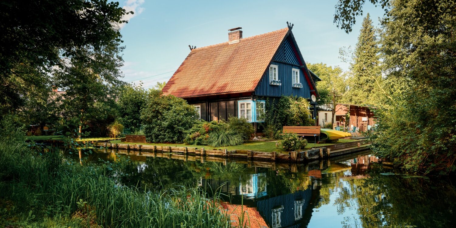 Ein schwarzes Haus mit rotem Dach steht in der Biege einer Flussschleife. Am Flussufer sind mehrere Kanus aufgebockt. Der Himmel über allem ist blau.