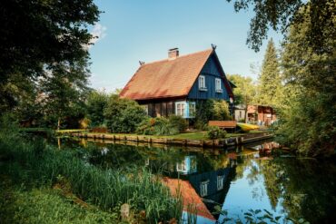Ein schwarzes Haus mit rotem Dach steht in der Biege einer Flussschleife. Am Flussufer sind mehrere Kanus aufgebockt. Der Himmel über allem ist blau.