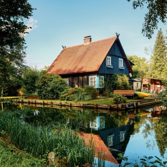 Ein schwarzes Haus mit rotem Dach steht in der Biege einer Flussschleife. Am Flussufer sind mehrere Kanus aufgebockt. Der Himmel über allem ist blau.