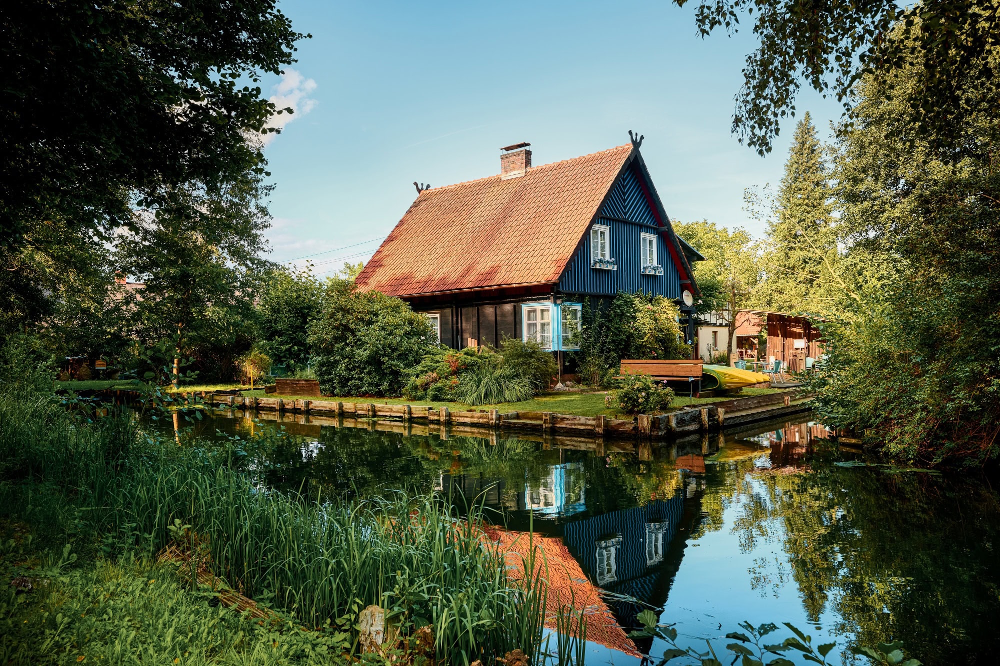 Ein schwarzes Haus mit rotem Dach steht in der Biege einer Flussschleife. Am Flussufer sind mehrere Kanus aufgebockt. Der Himmel über allem ist blau.