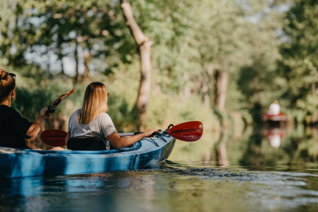 Zwei junge Frauen paddeln in einem Kanu durch einen kleinen Waldfluss.