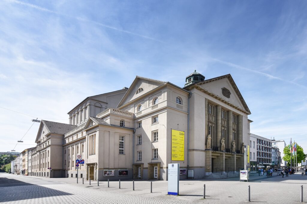 A white neoclassical building on the corner of a pedestrian zone, with a blue sky above it.