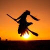 The silhouette of a young woman jumping into the air with a broom between her legs, against the setting sun. It looks as though she is flying on the broom.