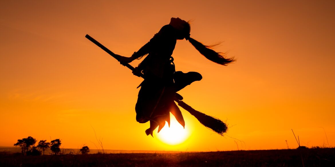 The silhouette of a young woman jumping into the air with a broom between her legs, against the setting sun. It looks as though she is flying on the broom.